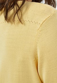 Gul stickad tröja med en slät yta, med dekorativa hål längs axelsömmen och rund halsringning.