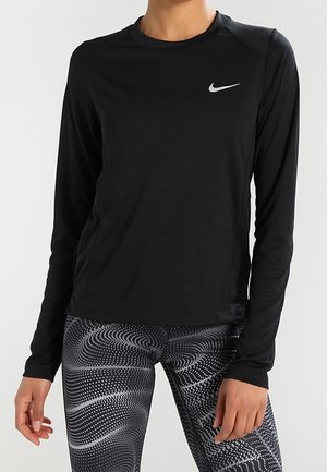 Zwart sportshirt met lange mouwen en een klein wit Nike-logo, gecombineerd met zwarte en witte leggings met patroon, gedragen door een staande persoon.