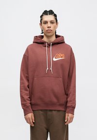 Bordová mikina s kapucňou vyrobená z bavlnenej zmesi, s predným kloktacím vreckom, sťahovacou kapucňou a oranžovým logom Nike na hrudi.