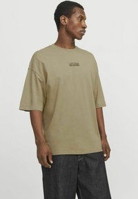 Camiseta de gran tamaño en verde oliva hecha de algodón suave, con un pequeño texto impreso en negro, combinada con pantalones de mezclilla oscuros.