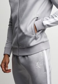 Tuta sportiva grigia composta da giacca con zip e tasche laterali, strisce bianche decorative e logo ricamato sui pantaloni. Materiale morbido.