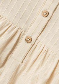 Beige textil med strukturerad yta, vertikala sömmar, rynkdetaljer och två runda träknappar med fyrhålsstygn.