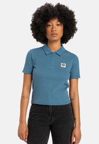 Un polo ajusté à manches courtes de couleur bleu atténué, présentant une texture côtelée et un petit logo brodé sur la poitrine gauche.