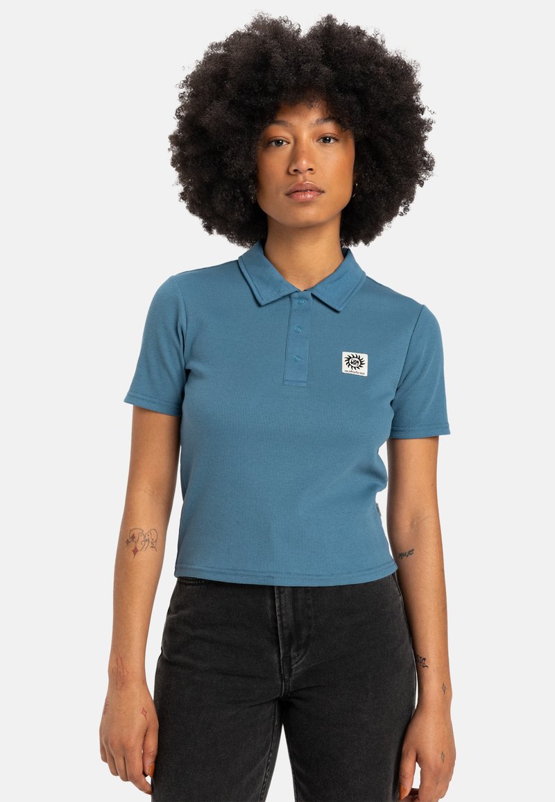 Un polo ajusté à manches courtes de couleur bleu atténué, présentant une texture côtelée et un petit logo brodé sur la poitrine gauche.