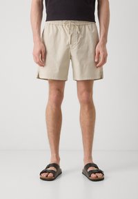 Pantaloni scurți bej din bumbac cu un tiv elastic și șnur. Purtați cu sandale negre, care au tălpi plate și curele late.