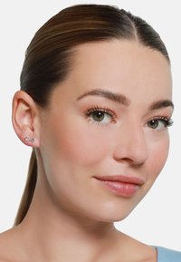 Silberohrringe in Form einer Schleife, mit einer polierten Oberfläche und einem minimalistischen Design, die am Ohr eines Modells präsentiert werden.
