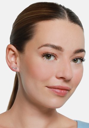 Silberohrringe in Form einer Schleife, mit einer polierten Oberfläche und einem minimalistischen Design, die am Ohr eines Modells präsentiert werden.