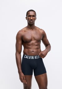 Fekete boxer alsónadrág széles fehér elasztikus derékpánttal, amelyen a "CALVIN KLEIN" logó található. Simán szőtt anyag és testhezálló kialakítás, napi viseletre alkalmas.