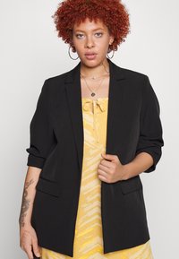 Blazer preto com ombros estruturados e frente aberta, apresentando dois bolsos frontais. Usado sobre um vestido amarelo estampado com detalhe de laço.