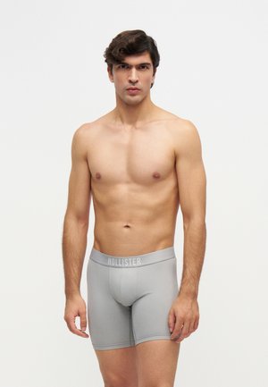 Skjorteløs ung mand iført lysegrå Hollister boxer, står foran en ensfarvet hvid baggrund.