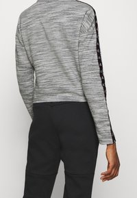 Maglione grigio a maniche lunghe, tagliato corto, con motivo testurizzato. Presenta strisce nere laterali con un dettaglio del logo. Abbinato a pantaloni neri.
