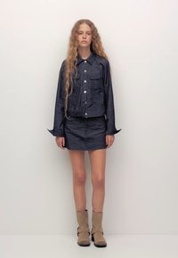 Giubbotto di jeans scuro con due tasche frontali, abbinato a una gonna coordinata. L'outfit presenta un design corto e dettagli con maniche svasate.