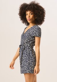 Navy dress with a wrap design, short sleeves, white spotted pattern, and a tie belt at the waist. Smooth fabric texture.