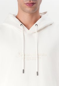 Weißer Kapuzenpullover aus weichem Stoff mit Kordelzug. Verfügt über gesticktes "Guess Jeans"-Logo und graue Metallelemente an den Kordeln.