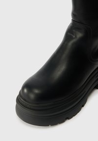 Bota ankle de couro preto com bico arredondado, sola de borracha elevada e detalhes de costura visíveis. A vista em close destaca a textura suave.
