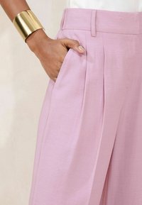 Pantaloni plissettati rosa chiaro con una texture liscia, dotati di tasche laterali. Un bracciale a manica dorata è indossato al polso.