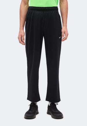 Persona che indossa pantaloni sportivi neri Nike, scarpe da ginnastica nere, calzini bianchi e una maglietta verde brillante, in piedi davanti a uno sfondo semplice.