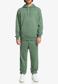 Zelená mikina s kapucňou a zodpovedajúce jogger nohavice, vyrobené z mäkkej látky. Mikina má klokanskú kapsu a šnúrku; jogger nohavice majú detail s logom.