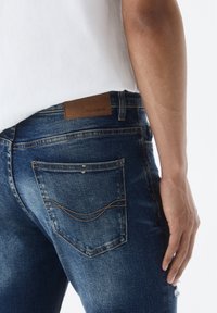 Jeansy dżinsowe w średnim niebieskim odcieniu z delikatnym blaknięciem. Posiadają skórzaną naszywkę z napisem "PULL&BEAR", widoczne przeszycia oraz detale z tylnych kieszeni.