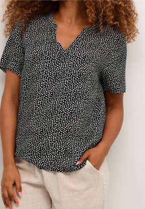 Mujer con blusa negra de manga corta con pequeñas formas blancas y pantalones beige claro, una mano en el bolsillo y cabello rizado visible.