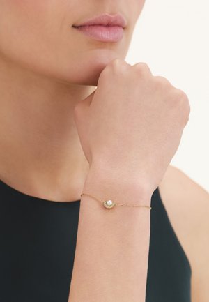Femme portant un délicat bracelet en or avec une petite perle et des diamants, reposant son menton sur son poing, sur un fond clair.