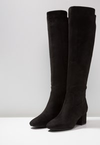 Bottes noires jusqu'au genou en daim souple. Dotées d'un design classique avec un talon épais et un bout arrondi. Texture lisse et finition sans coutures.