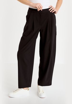 Kobieta w luźnych czarnych spodniach i białych trampkach, stojąca z jedną ręką na biodrze, a drugą poprawiająca pasek spodni.