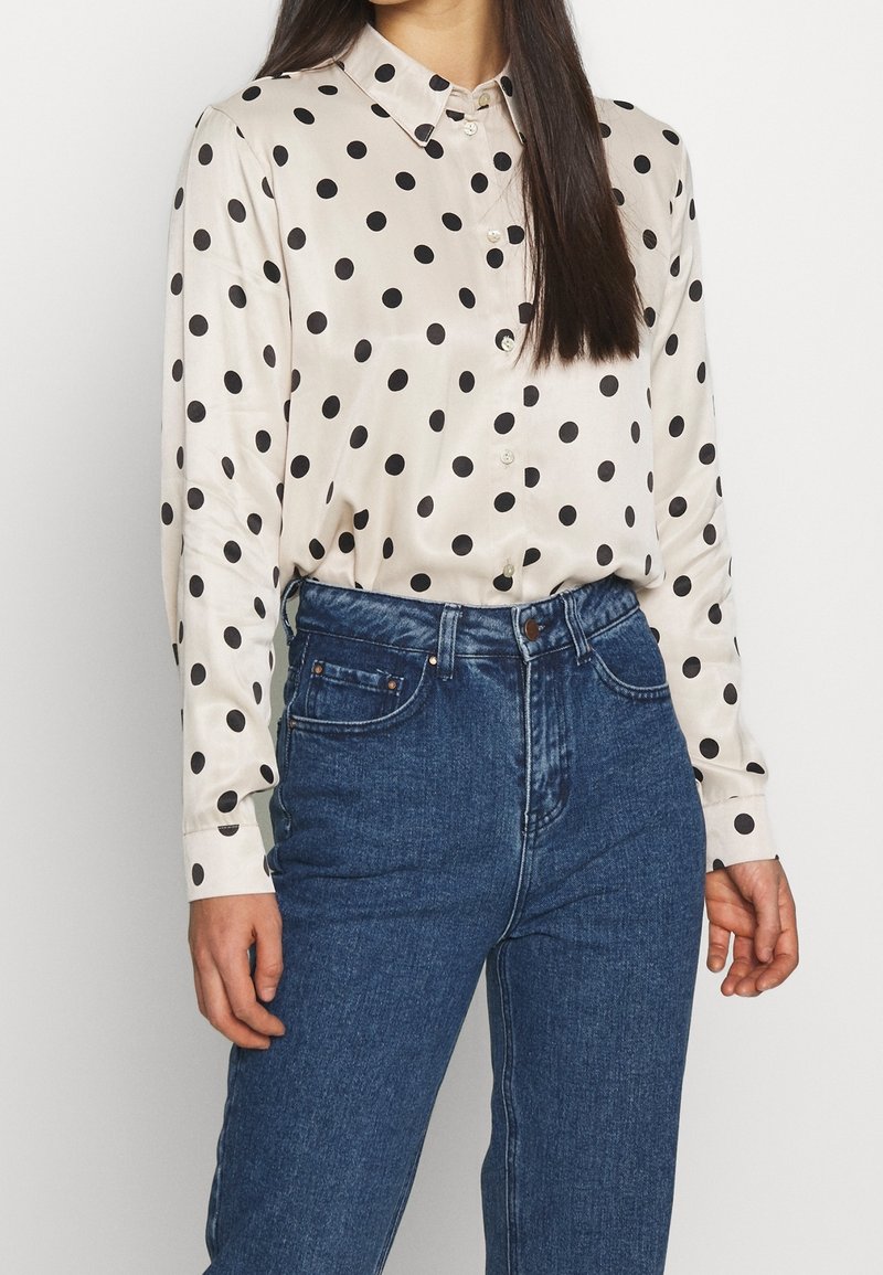 Camicetta in raso color crema conpois neri, colletto abbottonato e maniche lunghe. Abbinata a jeans in denim blu a vita alta.