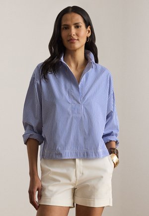 Frau trägt ein blau-weiß gestreiftes, langärmliges Hemd mit hochgekrempelten Ärmeln und beige Shorts, steht vor einem schlichten Hintergrund.