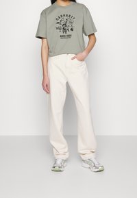 Vaaleanvihreä lyhythihainen t-paita mustalla graafisella kuviolla, yhdistetty off-white leveälahkeisiin housuihin ja harmaisiin lenkkareihin, joissa on vihreitä yksityiskohtia.