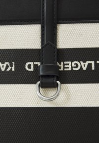 Détail d'un sac en toile rayé noir et crème, avec une bandoulière en cuir équipée d'un anneau en métal argenté et un logo embossé.