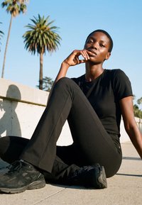 Lühikeste juustega naine mustas riietuses istub väljas kõnniteel betoonseina lähedal, taustal palmid.