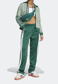 Grön träningsdräkt med vita sidostripes, tillverkad av slätt tyg. Matchas med en grön midjeväska och retroinspirerade sportskor med ränder.