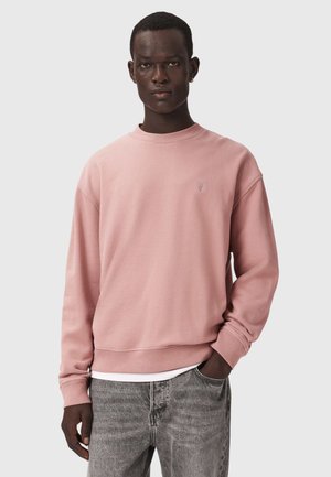 Jonge man die een roze trui met ronde hals draagt over een wit overhemd en een grijze spijkerbroek, met één hand in de zak tegen een effen achtergrond.