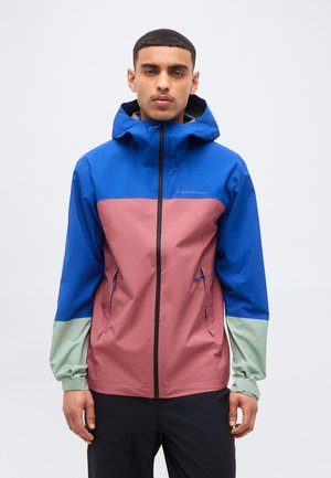 Noor mees kannab värvilise kujundusega jopet sinise kapuutsi ja õlgadega, tolmroosa kere ja heleroheliste varrukaservadega, seisab valge taustaga.