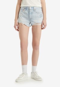 Pantalones cortos de mezclilla azul claro con dobladillo deshilachado, un ajuste relajado, cinco bolsillos y costuras en contraste. Combinados con zapatillas blancas y calcetas.