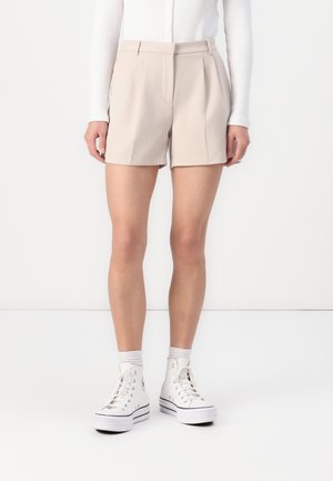 Mujer con pantalones cortos beige a medida, una camiseta blanca de manga larga acanalada, calcetines blancos y zapatillas blancas de caña alta, de pie contra un fondo blanco.