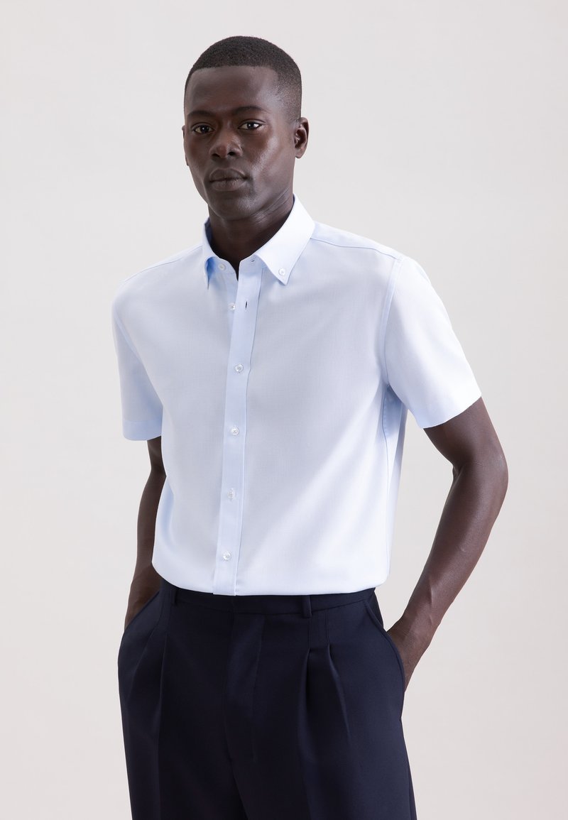 Homme portant une chemise à manches courtes bleu clair rentrée dans un pantalon plissé bleu marine foncé, debout les mains dans les poches.