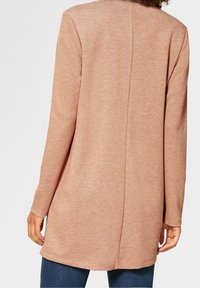 Pitkä, tyköistuva takki pehmeästä, tekstuurilla varustetusta kankaasta, vaaleanpunainen väri, jossa on suora selkä näkyvillä sauma-yksityiskohdilla ja pitkät hihat.