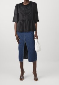 Blusa nera semi-trasparente con dettagli arricciati, abbinata a una gonna di jeans di lunghezza media con uno spacco frontale. La modella tiene una borsa bianca.