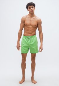 Νεαρός άνδρας όρθιος, ξυπόλυτος, φορά φωτεινά πράσινα μαγιό, πάνω σε απλό λευκό φόντο.