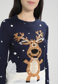 Granatowy sweter dziany z motywem renifera, płatków śniegu i zaśnieżonego krajobrazu u dołu. Miękka faktura, okrągły dekolt.