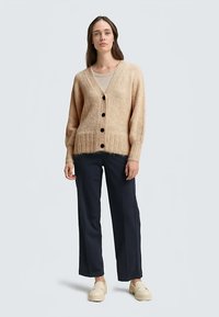 Beige wollen vest met een V-hals, lange mouwen en een geribbelde zoom. Heeft donkere knopen aan de voorkant, gecombineerd met marineblauwe rechte broek.