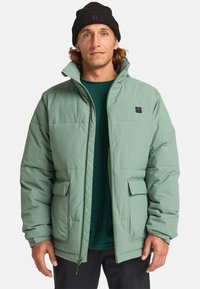 Giacca imbottita verde chiaro con colletto alto, due tasche frontali, patch logo nero e cerniera, indossata sopra una camicia verde scuro.
