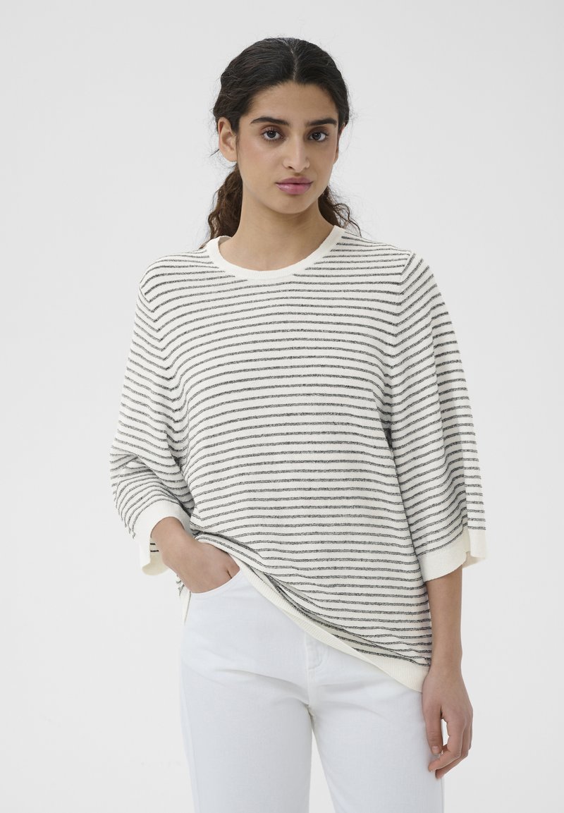 Femme aux cheveux foncés portant un pull ample rayé blanc et noir et un pantalon blanc, debout, une main dans la poche, sur un fond uni.