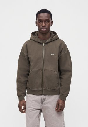 Uomo che indossa una felpa con cappuccio verde oliva con zip e tasche frontali e jeans beige, in piedi davanti a uno sfondo chiaro e uniforme.