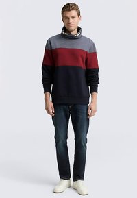 Stickad tröja i grått, rött och marinblått, gjord av mjukt tyg. Har en hög krage med knappradie. Bärs med mörka jeans och sneakers.