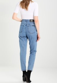 Džíny s vysokým pasem modré barvy vyrobené z denimu, s dvěma zadními kapsami a štítkem na pasu, stylizované s černými kotníkovými botami.