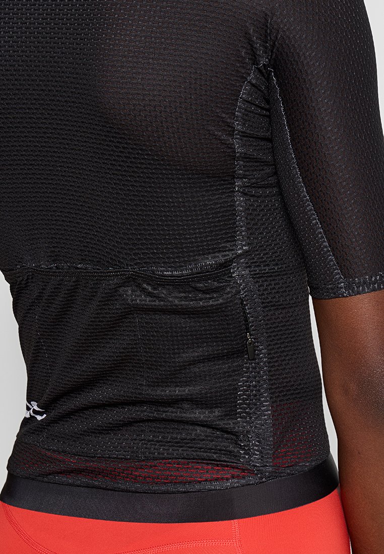 Schwarzes, strukturiertes Sportoberteil mit kurzen Ärmeln und einer seitlichen Reißverschlusstasche, getragen mit roten Sporthosen.