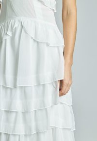 Weißes, gestuftes Kleid mit Rüschenschichten, leichtem Stoff, weicher Textur und tailliertem Oberteil, das eine Hand nahe dem unteren Saum zeigt.
